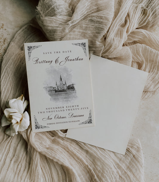 New Orleans wedding save the date with a watercolor image of the St. Louis Cathedral on parchment paper and black writing. Lovely fleur de lis corner ornamental decorations grace the corners.